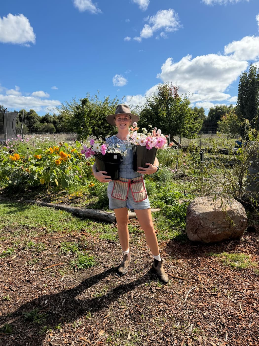 Grace of Feel Good Farm Flowers carrying two buckets of cut dahlias and cosmos, with WildSoil mulch through the beds behind her